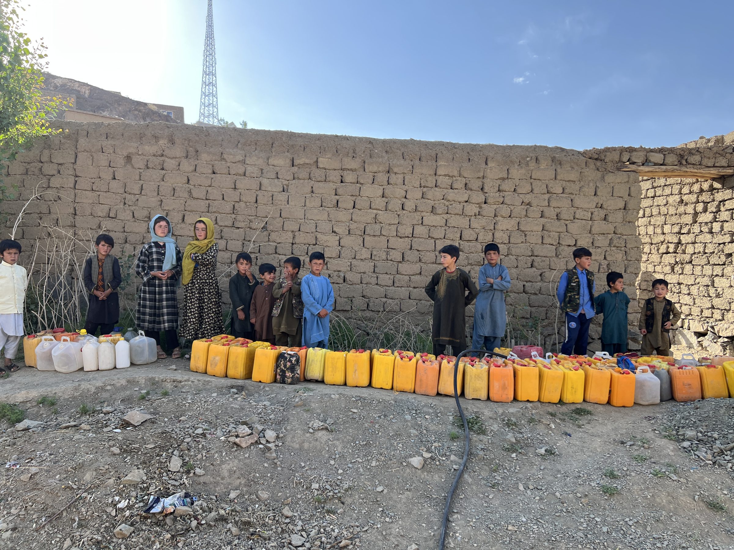 A long line of children beside water jerrycans along a mud-brick wall in Ghor